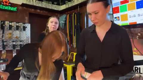 Media: Video of three women in a salon: one cutting hair, one styling, and another observing. Background features colorful artwork, a bar, and a large screen displaying digital content.