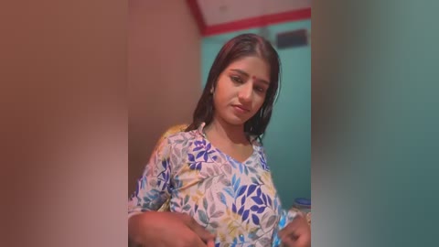 Media: A video of a young South Asian woman with straight black hair, wearing a colorful floral blouse, standing in a simple room with teal walls and a red-framed window.