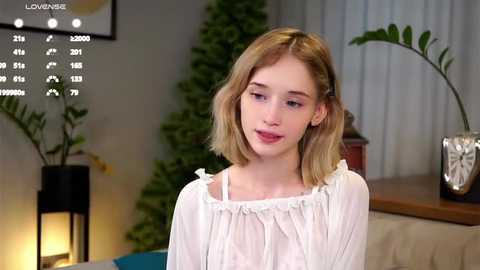 Media: Video of a young, fair-skinned woman with shoulder-length, wavy blonde hair, wearing a white, off-shoulder blouse, smiling gently, indoors with a calendar, potted plants, and a table.