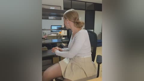 Media: A video of a blonde woman in a white sweater and beige skirt working at a modern office desk with a computer monitor and files.