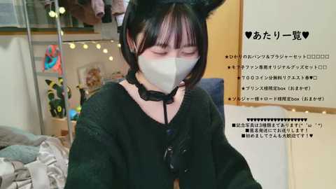 Media: Video of an East Asian woman with short black hair, wearing a black mask and sweater, seated in a cozy room with string lights, a bed, and Japanese text.