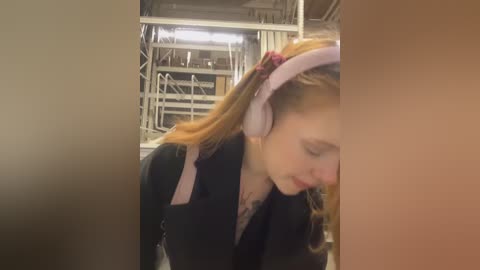 Media: Video of a young woman with long, wavy red hair, wearing pink headphones, a black blazer, and a white shirt, looking down in a dimly lit room with metal scaffolding and shelves in the background.