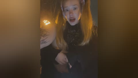 Media: A video captures a young woman with fair skin and long, straight blonde hair, wearing a black sweater, with a distressed expression, illuminated by a dim, warm light. She is standing in a dark, possibly misty, background.