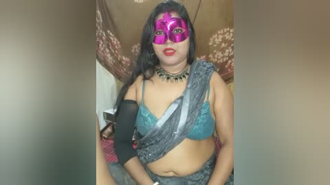 Media: Video of a South Asian woman with medium brown skin, wearing a turquoise bra, grey sari, and a pink Venetian mask, standing in a dimly lit room with floral-patterned curtains.