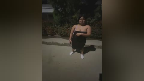 Media: Video of a plus-size Latina woman with dark hair, wearing a strapless black dress and white shoes, standing outdoors on a concrete path. Background features green foliage and a white building.