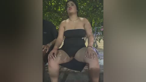 Media: Video of a plus-sized woman with light brown skin and black hair in a strapless black dress, sitting on a bench outdoors, surrounded by green foliage and a blurred man.