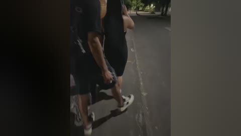 Media: Video of two men in dark clothing, one with a white shirt, the other in a black shirt, skateboarding on a deserted road, shadows and dim lighting creating a moody atmosphere.