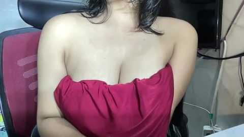 Media: A video of a woman with fair skin and shoulder-length dark hair, wearing a strapless red dress that reveals her cleavage, sitting on a red office chair. Background shows a cluttered office space with a computer monitor and wires.