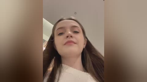 Media: Video of a young woman with long brown hair, light skin, and a slight smile, wearing a white sweater, captured from a low angle with blurred objects in the foreground.