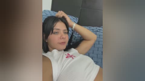 Media: A video of a young woman with light skin and long black hair, wearing a white ribbed tank top, lying on a bed with blue brick-patterned sheets, looking relaxed and slightly bored.