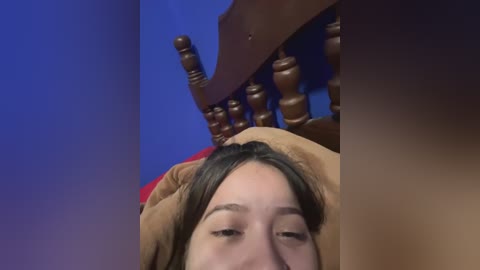 Media: A video captures a young woman with light skin and brown hair, lying on a wooden bed frame with a blue wall background, partially covered by a brown blanket.