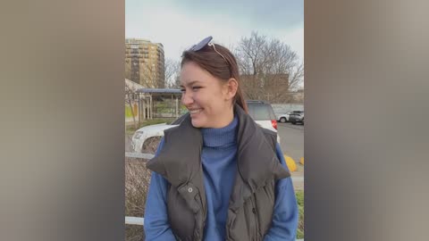 Media: Video of a smiling Caucasian woman with auburn hair, wearing a blue turtleneck under a black puffer vest, standing in a parking lot with modern buildings and cars in the background.