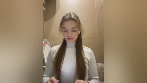 Media: Video of a young Caucasian woman with long, straight brown hair, wearing a white turtleneck sweater, seated in a beige-tiled bathroom, looking down at a small device, with a fan and a sink in the background.