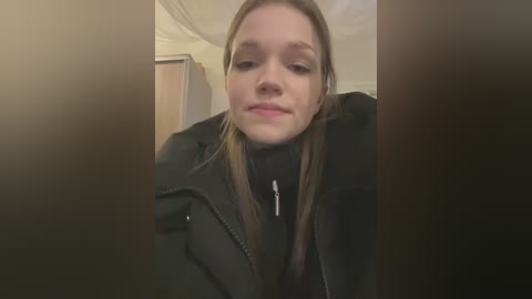 Media: Video of a young Caucasian woman with light skin, brown hair, and brown eyes, wearing a black jacket, leaning against a wall with a neutral expression.