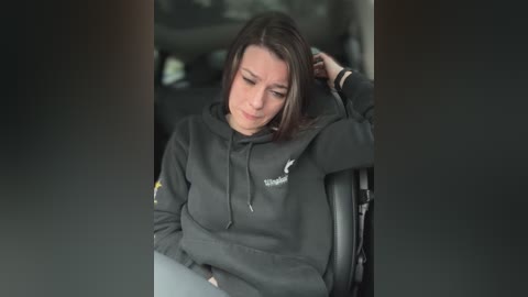 Media: A video shows a young woman with fair skin and straight brown hair, wearing a black Adidas hoodie, seated in a car, looking tired and leaning against the seat.