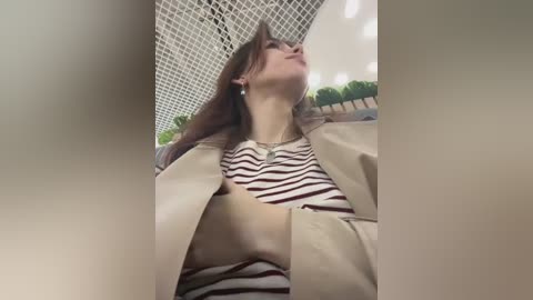 Media: Video of a young woman with long brown hair, wearing a beige leather jacket over a striped top, standing in a modern indoor space with white grid ceiling and green plants.