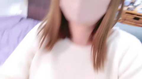 Media: Video of a woman with fair skin and shoulder-length light brown hair, wearing a white sweater, lying on a bed with lavender sheets. The background shows a blurred wooden dresser and a wall.