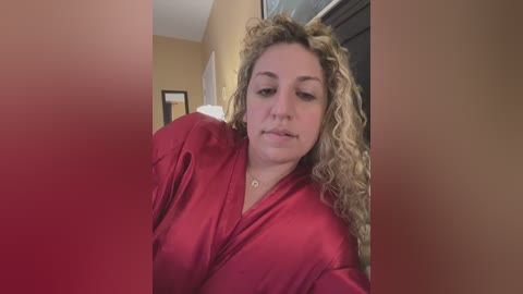 Media: A video of a blonde woman with curly hair, wearing a red satin robe, standing in a beige-walled room with a window and framed picture in the background.