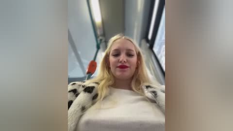 Media: Video of a blonde woman with fair skin and red lipstick, wearing a white sweater with black polka dots, looking relaxed on a subway train.