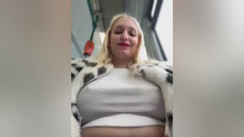 Media: Video of a blonde woman with fair skin, wearing a white crop top and a cow-patterned jacket, seated on a bus. She has a neutral expression and the background shows a bus interior.