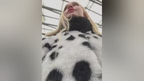 Media: A video captures a low-angle view of a woman with long blonde hair, wearing a fur coat resembling a panda with black and white patches. The background is a modern, glass-walled structure.