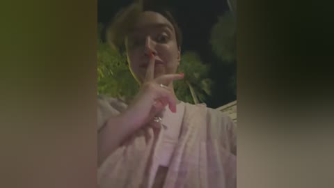 Media: Video of a woman with fair skin, dark hair, and red nails, holding her finger to her lips, shushing, against a blurred background of trees and a house at night.