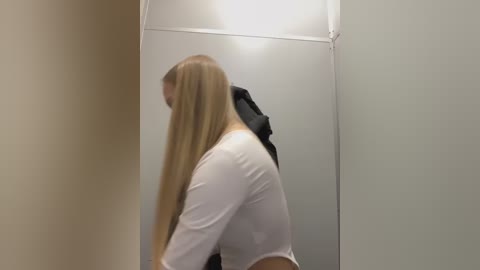 Media: A video captures a blonde woman with long hair in a white crop top, standing in a small, dimly lit bathroom stall. The background features a gray door and beige walls.