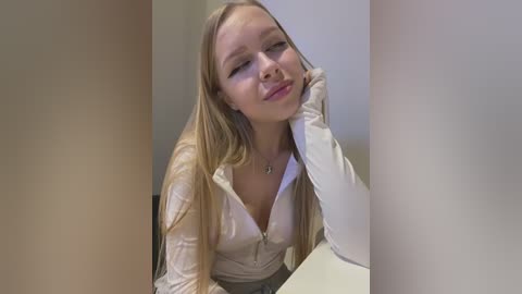 Media: Video of a young blonde woman with long hair, wearing a white blouse, leaning on her hand, smiling in a casual indoor setting.