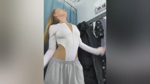 Media: Video of a slender, blonde woman in a white, high-cut bodysuit, and grey skirt, leaning against a clothing rack filled with black and white garments in a brightly lit, modern dressing room.