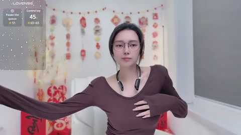 Media: Video of an Asian woman with dark hair, wearing glasses and a brown top, adjusting her headphones in a modern, white room with red and orange decorations.