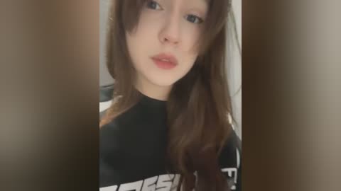 Media: A video of a young woman with long brown hair, fair skin, and a slight smile. She wears a black T-shirt with white text. The background is blurred.