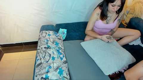 Media: Video of a tattooed woman in a purple tank top and jeans, sitting on a blue couch, reading a book, with a blue star-patterned blanket and a gray pillow beside her.