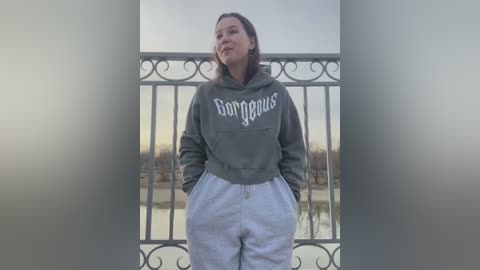 Media: Video of a young woman with straight, shoulder-length hair, wearing a grey \"Gorgeous\" hoodie and white sweatpants, standing on a balcony with a serene lake and autumn foliage in the background.