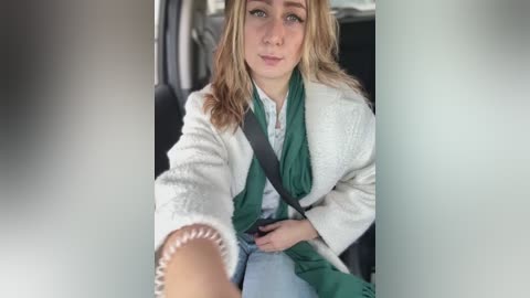 Media: Video of a young woman with fair skin, blonde hair, and freckles, wearing a white quilted coat, green scarf, and blue jeans, sitting in a car, seen from the front seat.