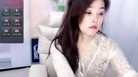 Media: Video of a young Asian woman with long, straight black hair, wearing a white lace dress, sitting in a white leather chair, looking at a digital clock displaying 3:19 PM, in a modern room.