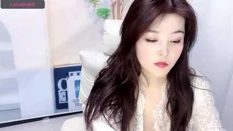 Media: Video of an East Asian woman with long, dark hair, fair skin, and red lipstick, wearing a white lace top, sitting indoors. Background features a white chair, potted plant, and blue box.