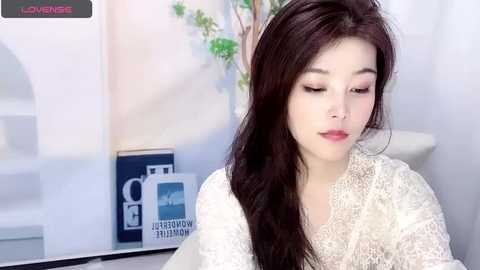 Media: A video of a young East Asian woman with long, dark hair and fair skin, wearing a white lace robe, sitting indoors. The background features a white wall with a potted plant and a blue informational sign.