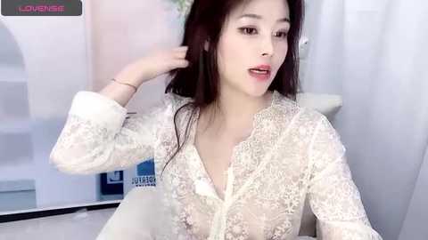 Media: Video of an East Asian woman with fair skin and long black hair, wearing a sheer, lace-up white blouse, seated in a modern, minimalist room.