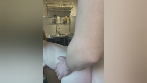 Media: Video of a naked person, likely female, standing in a kitchen with pale skin, holding their thighs. The background features a stainless steel sink, utensils, and a wall-mounted shelf.