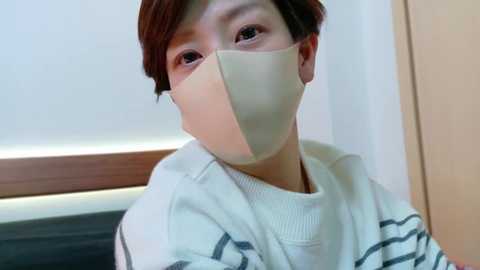 Media: Video of an East Asian person with short brown hair, wearing a beige surgical mask and a white, striped sweater. They are indoors with a wooden panel and white wall in the background.