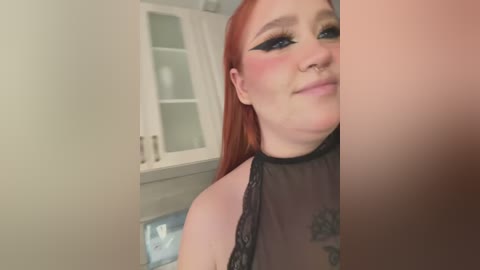 Media: A video of a fair-skinned woman with long, straight red hair, wearing black lace top, heavy black eyeliner, and a septum piercing, smiling softly in a modern kitchen with white cabinets and glass doors.