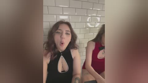 Media: Video of two women, one with long, wavy brown hair, wearing a black halter top with a keyhole cutout, and another with long brown hair, in a red top, both looking towards the camera in a bathroom with white tiled walls.