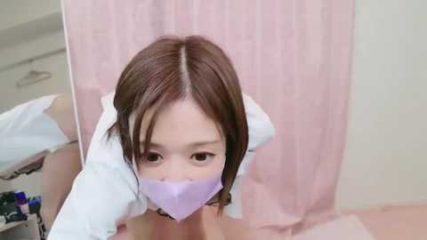 Media: Video of a young East Asian woman with straight brown hair, wearing a white shirt and a purple face mask, lying on a bed with pink sheets and a white curtain in the background.