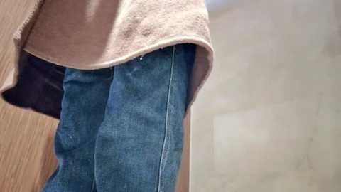 Media: Video of a person's lower body, showing blue jeans and a beige towel draped over the back, against a blurred, light-colored background.