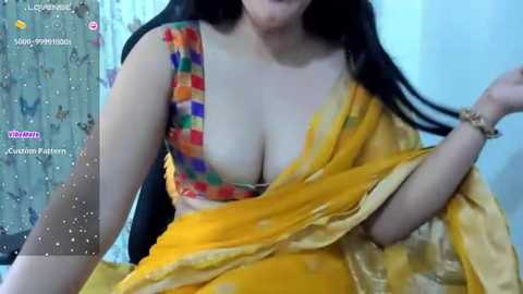 Media: Video of a fair-skinned woman with long black hair, wearing a yellow saree and a colorful bralette, indoors with a patterned curtain background.