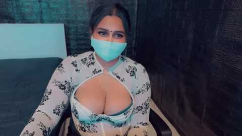 Media: Video of a South Asian woman with fair skin, wearing a white floral-patterned top that reveals her ample cleavage, a turquoise face mask, and dark hair tied back. She sits on a bed in a dimly lit room with dark brick walls.