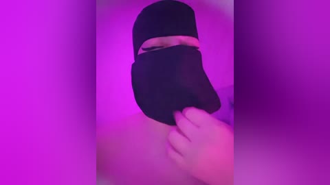 Media: Video of a person wearing a black face mask, with hands covering part of the face. Background is a gradient of purple hues, creating a moody atmosphere.