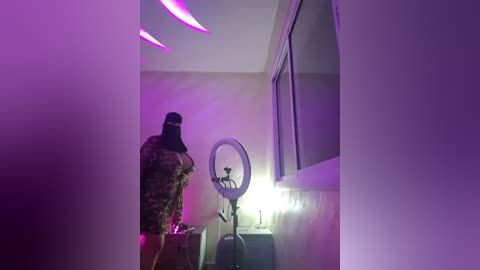 Media: Video of a dimly lit room with a person wearing a camouflage jacket and hood, standing near a round light and a window, under purple LED lights.