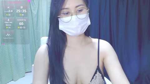 Media: Video of an East Asian woman with long black hair, wearing a white surgical mask, glasses, and a black lace bra, sitting in a hospital room with a green curtain and a digital clock showing 25:35.