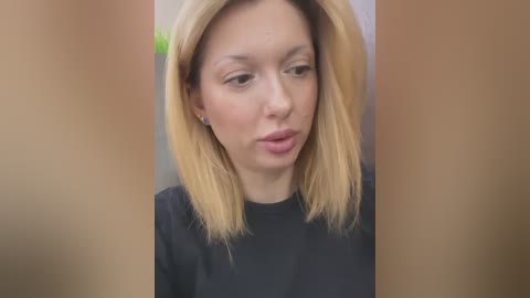 Media: Video of a light-skinned woman with blonde shoulder-length hair, wearing a black top, looking thoughtful against a blurred, neutral background.